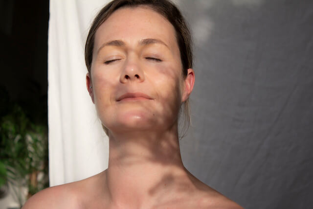 Benefits of Manuka Oil image with a woman basking in sunlight with clear skin and the shadows of Manuka plant on her face.