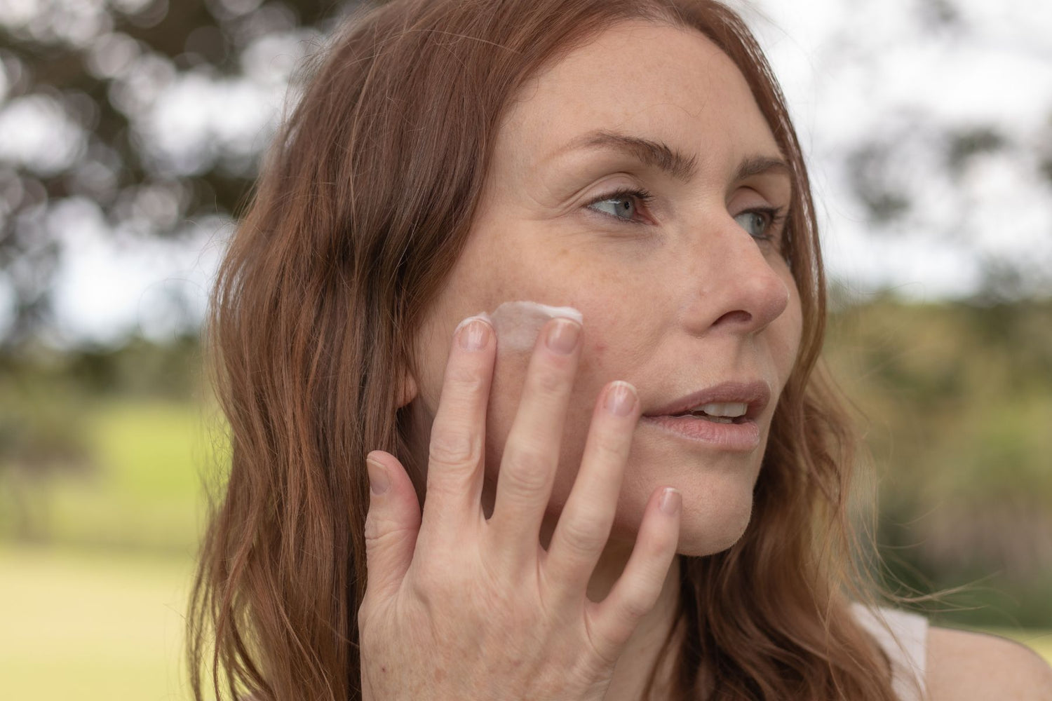 A woman in the midst of nature, tenderly applying a hydrating cream onto her face, surrounded by lush greenery and serene natural beauty, embracing the nourishing benefits of skincare while immersing herself in the tranquility of the outdoors